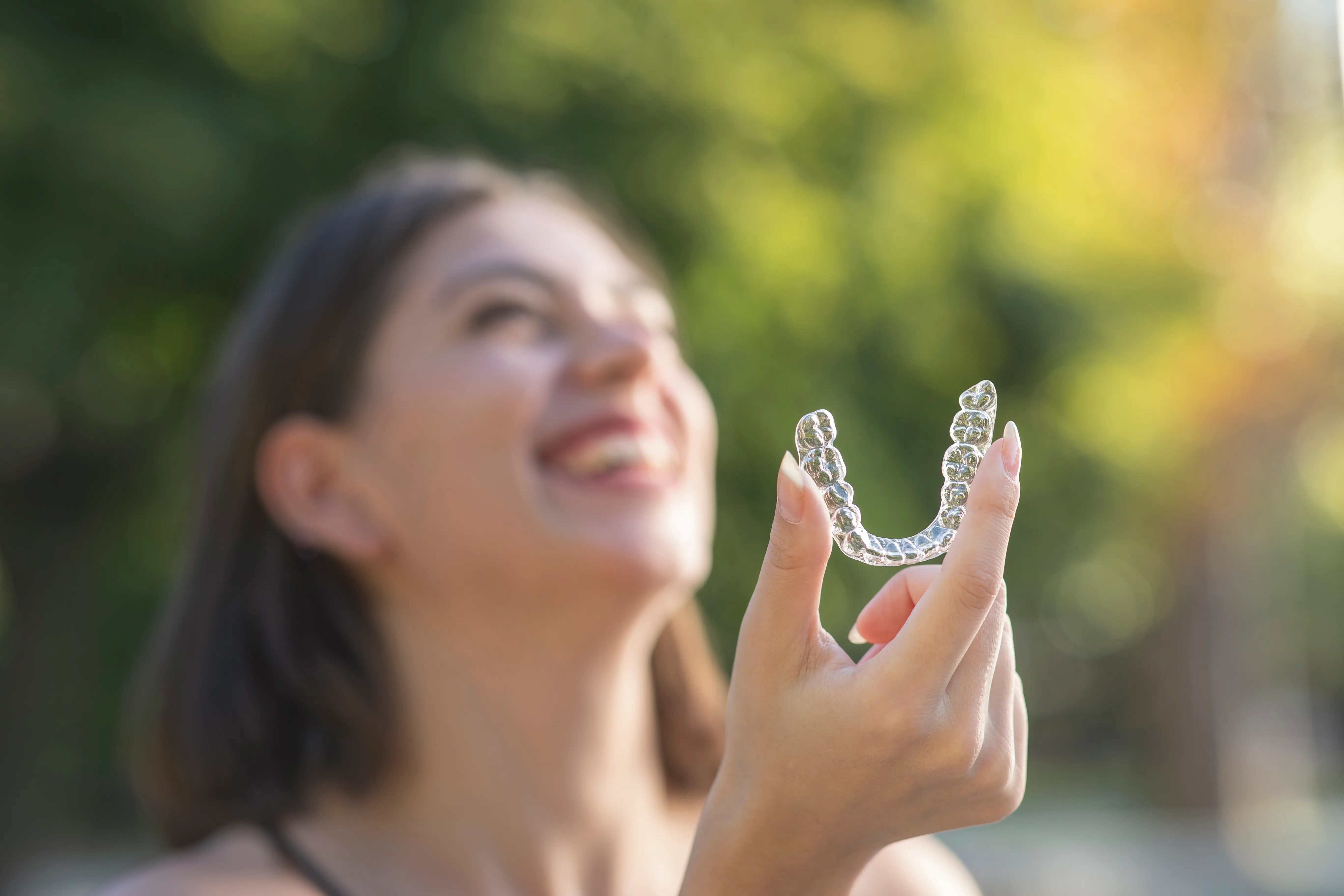 Smiling woman in outdoor setting, holding a clear aligner.